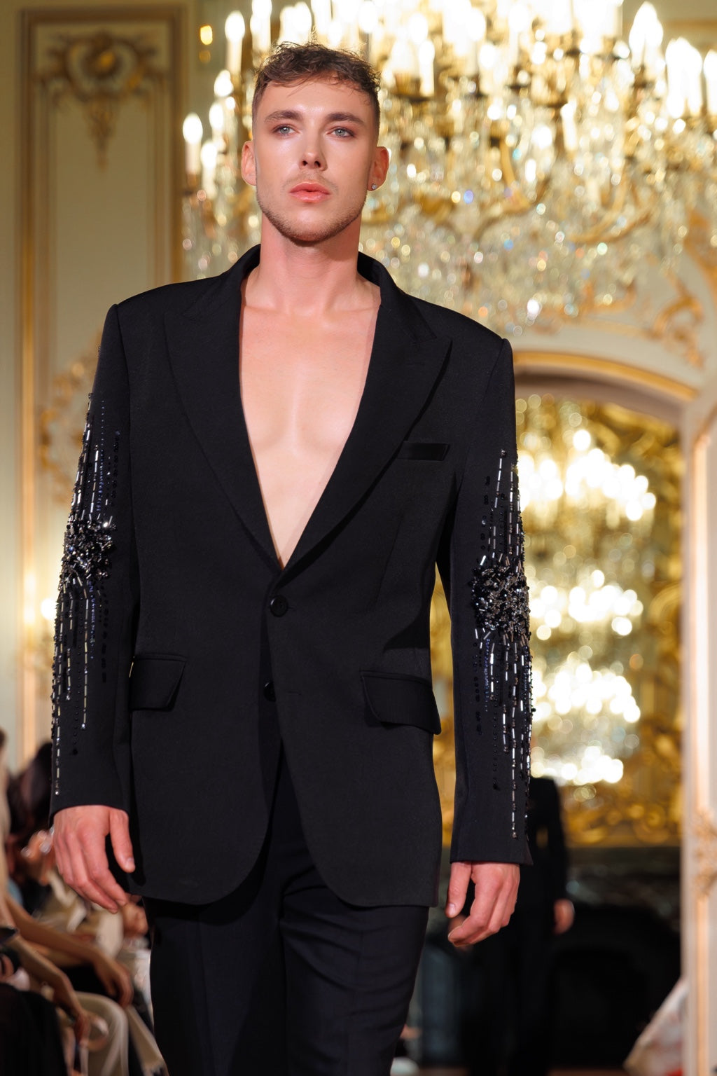 Clément Rudnicki on the runway, wearing an embroidered black jacket beneath chandeliers.